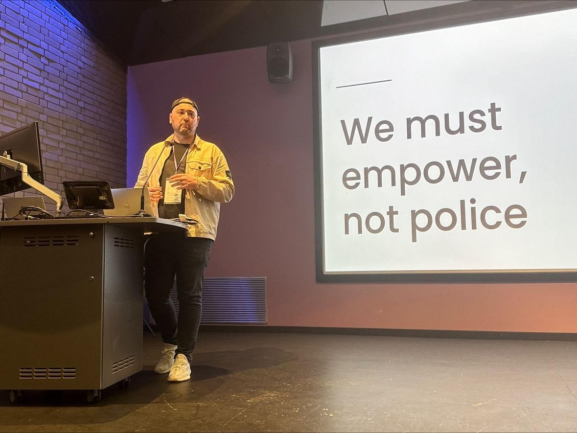 Shaun wearing black jeans and Tshirt with a beige jacket and his cap on backwards standing behind a lectern in front of a big screen that says we must empower, not police.