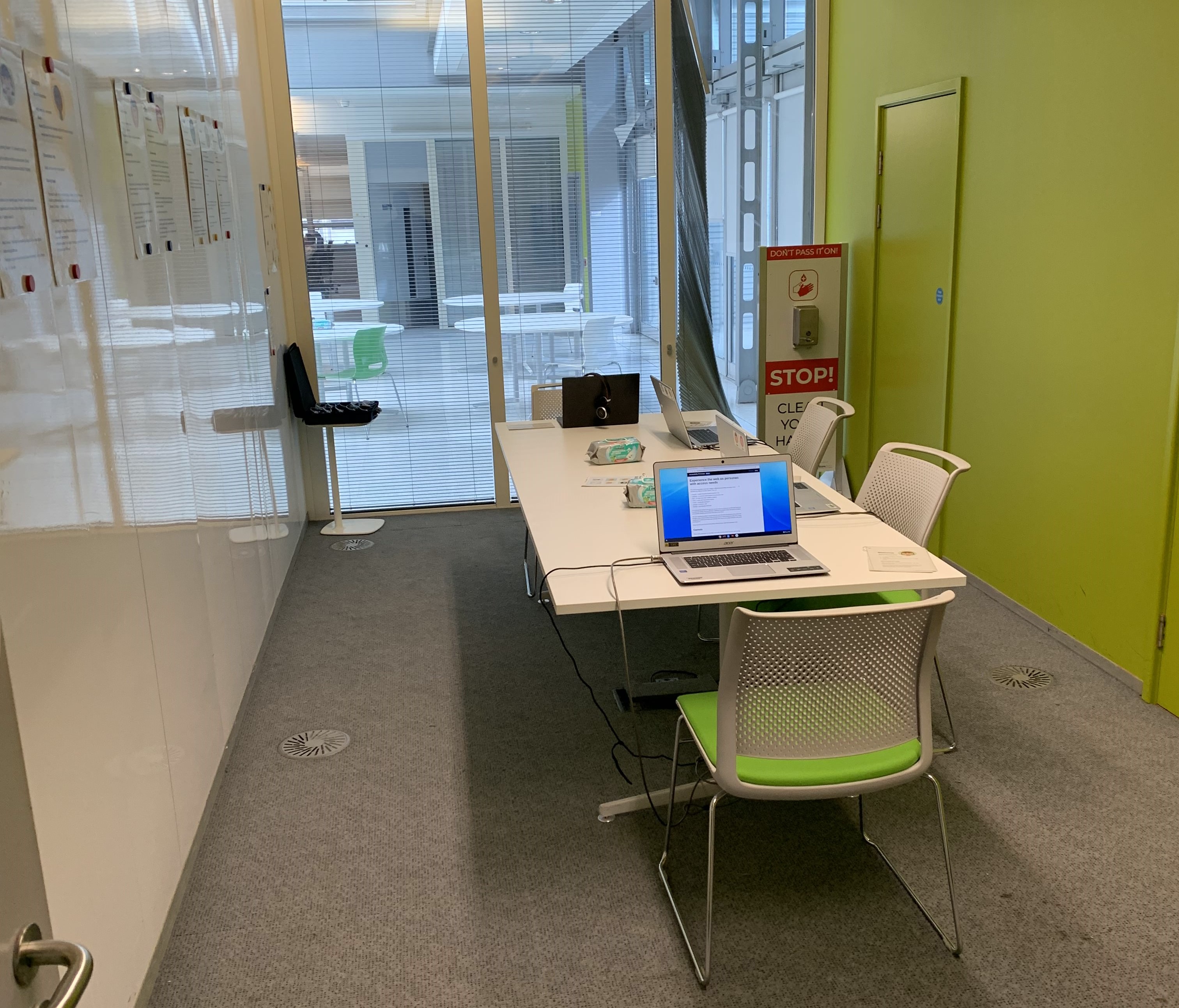 A large table with 4 seats in the middle of a small room. There are 4 laptops on the table, a box with simulation glasses is at the back of the room and there are some persona posters on the wall